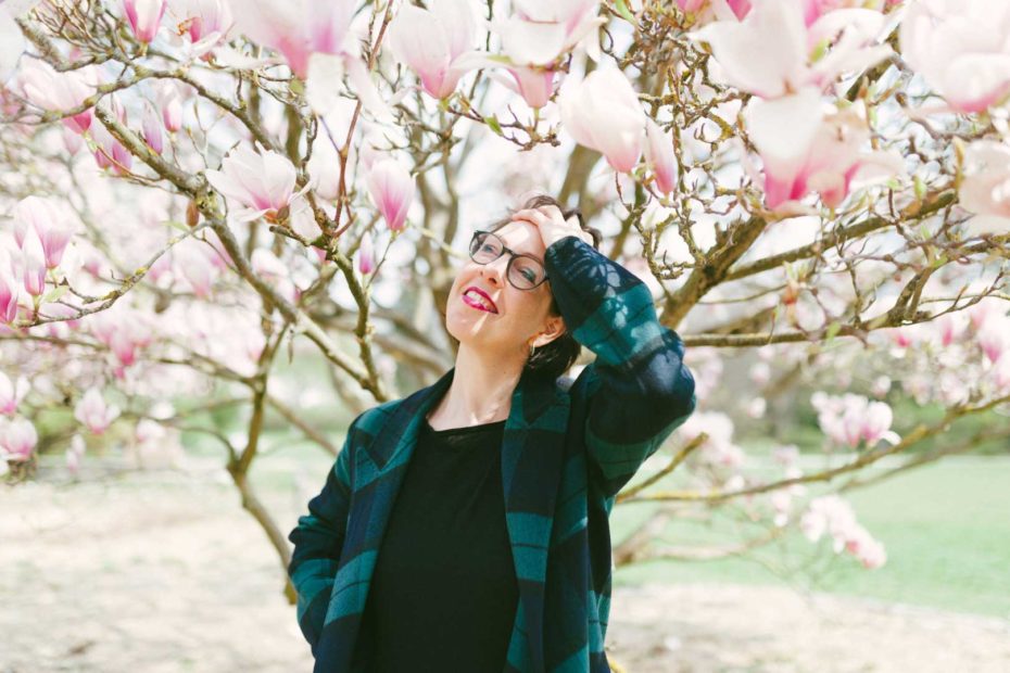 Stilberaterin Caro, die sich vor einem wunderschönen Blütenbaum im Sonnenschein in die Haare fasst und lächelt.